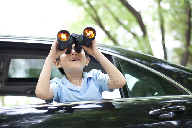Cheerful Chinese boy leaning out of car window and looking with binoculars — Stock Photo