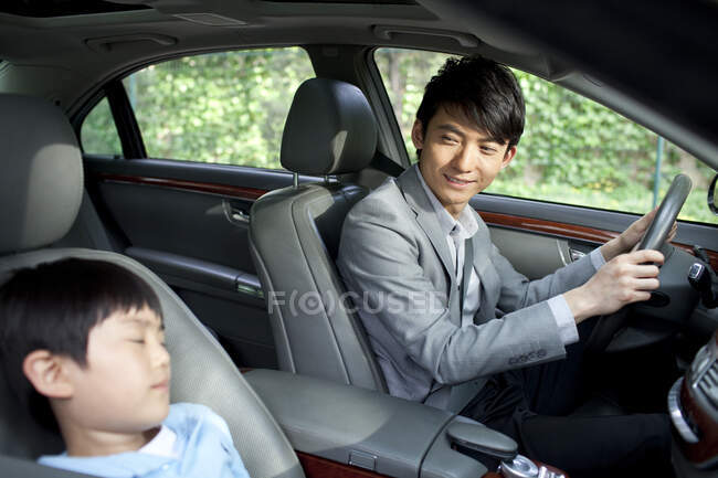Alegre padre chino conduciendo con su hijo durmiendo en el asiento delantero - foto de stock