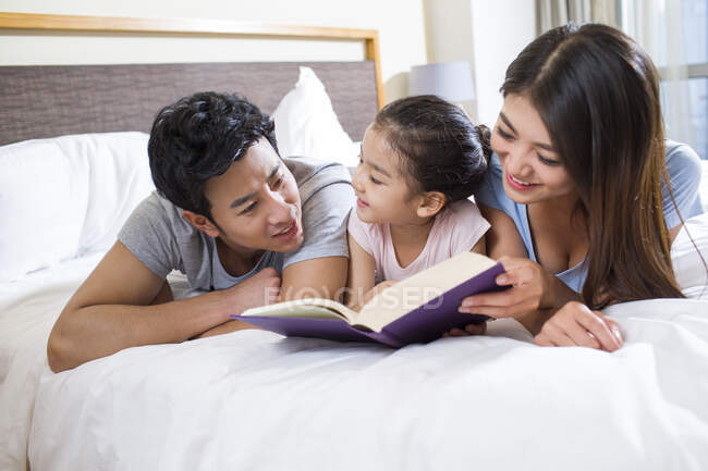 Веселая молодая китайская семья читает книгу на кровати — стоковое фото
