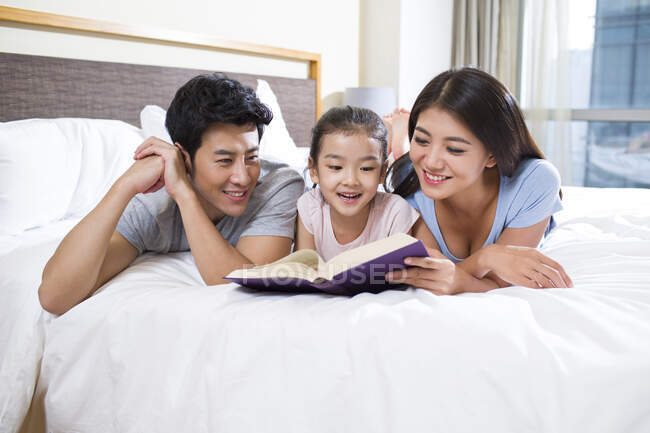 Joven familia china alegre leyendo un libro en la cama - foto de stock