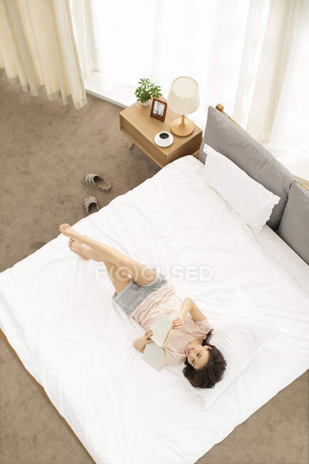 Glückliche junge Chinesin liest Buch auf dem Bett — Stockfoto