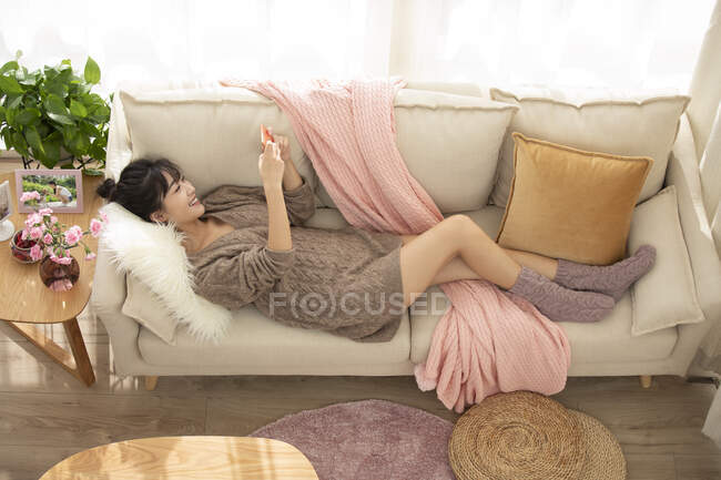 Joven mujer china jugando juegos móviles en el sofá - foto de stock