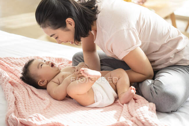 Joven madre china sonriendo y jugando con el bebé en el sofá - foto de stock