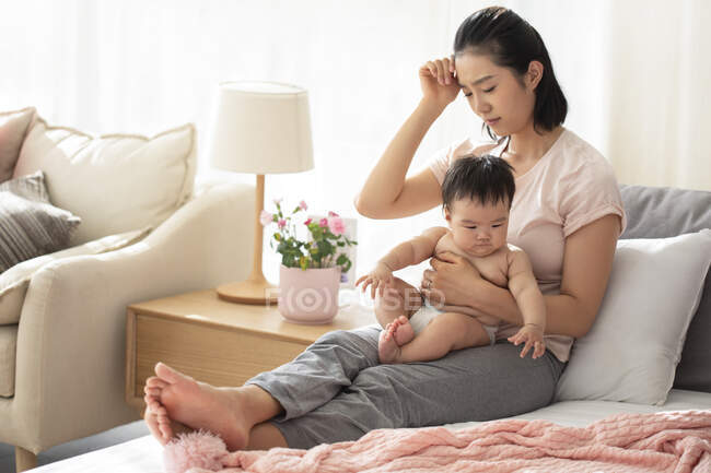 Втомлена мама тримає дитину, сидячи на дивані з рукою на голові — стокове фото