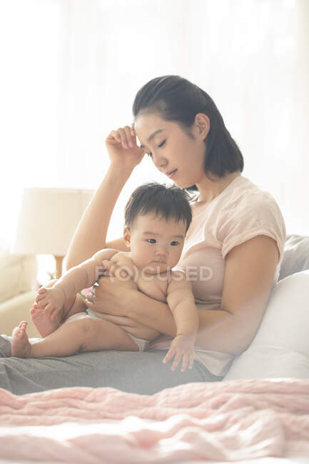 Madre cansada sosteniendo al bebé mientras está sentada en el sofá con la mano por la cabeza - foto de stock