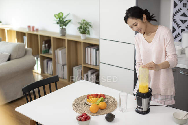 Joven mujer china sonriendo y haciendo jugo fresco en casa - foto de stock