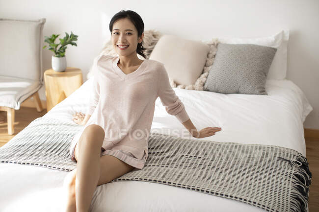 Счастливая китаянка, сидящая на кровати и улыбающаяся — стоковое фото