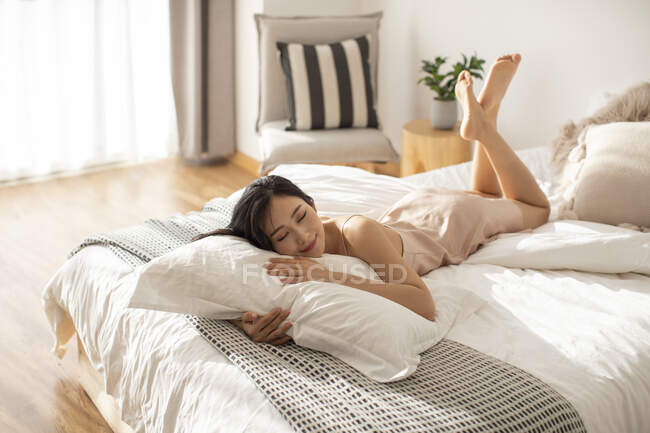 Feliz joven china acostada con almohada en la cama y sonriendo con los ojos cerrados - foto de stock