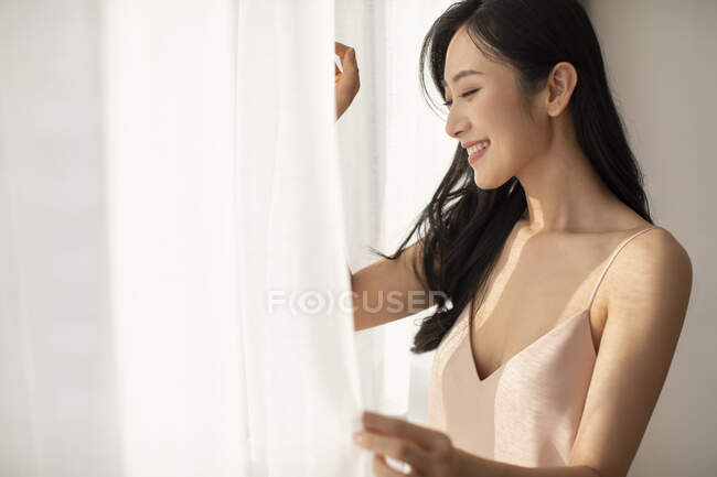Jovem chinesa feliz de pé com cortinas na janela e sorrindo — Fotografia de Stock
