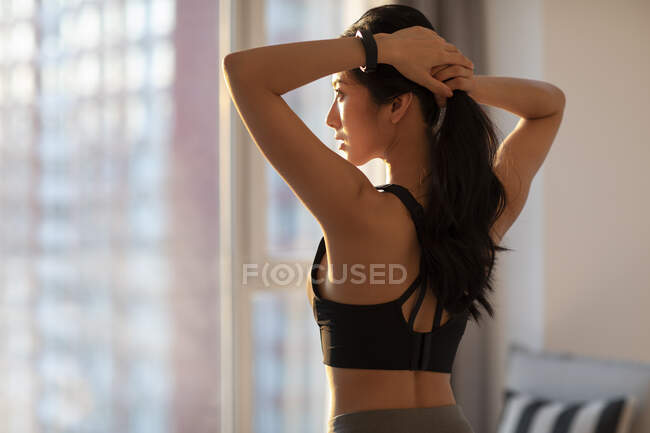 Joven mujer china en ropa deportiva atando el cabello a la luz del sol de la mañana - foto de stock