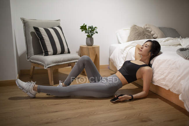 Joven mujer china descansando en el suelo por la cama después del entrenamiento - foto de stock