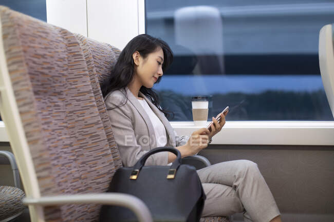 Femme assise dans un train à grande vitesse avec tasse de café et utilisant un smartphone — Photo de stock