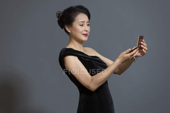 Mujer china madura en vestido negro mirando espejo de bolsillo - foto de stock
