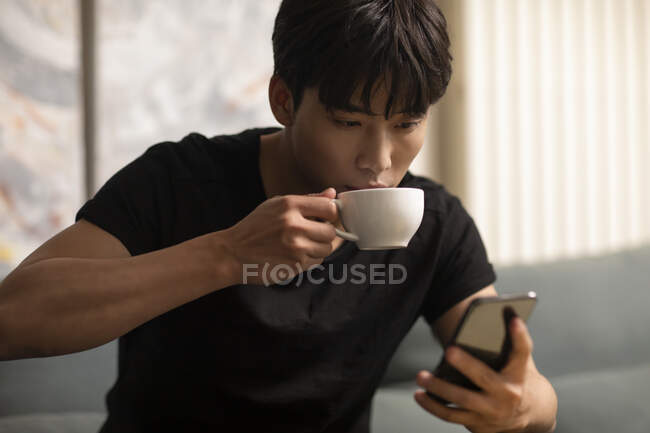 Young chinese man drinking coffee from cup and looking at smartphone screen — Stock Photo