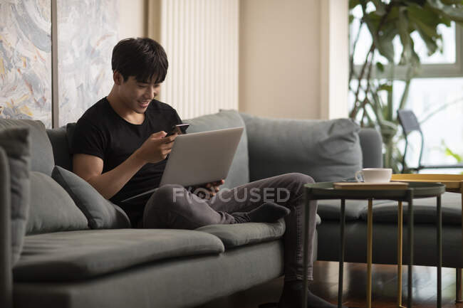 Улыбающийся китаец смотрит на экран смартфона, держа в руках ноутбук — стоковое фото
