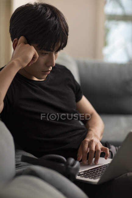 Молодий китайський чоловік використовує ноутбук, сидячи на дивані — стокове фото