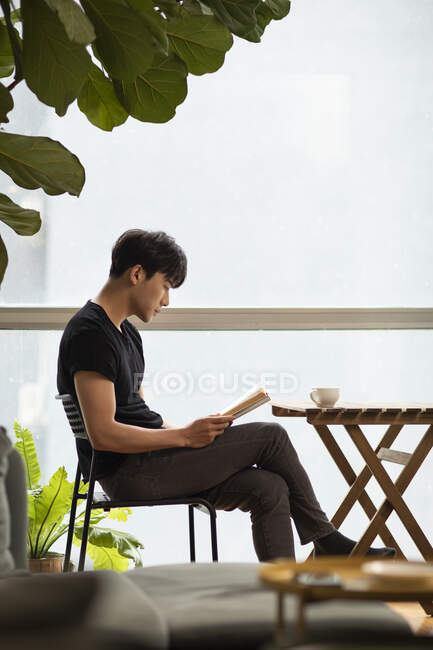Young chinese man reading book while sitting at table with coffee cup — Stock Photo