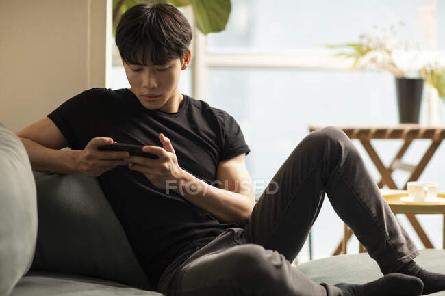 Молодий китайський чоловік використовує смартфон, сидячи на дивані — стокове фото