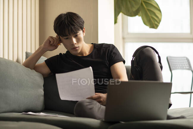 Young chinese man looking at paper sheet while sitting on couch with laptop — Stock Photo