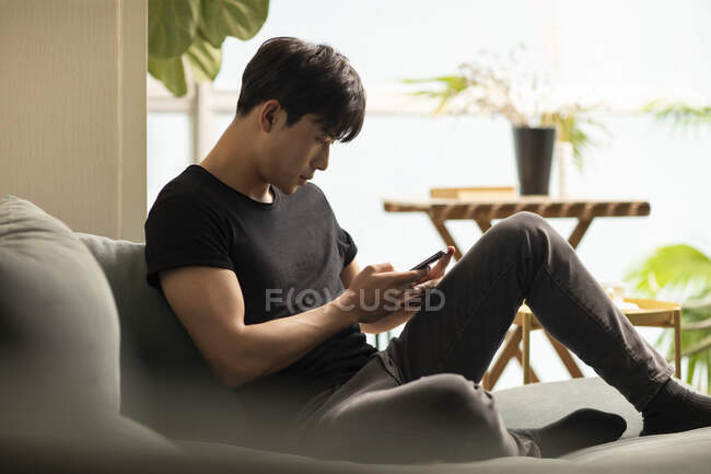 Junger Chinese benutzt Smartphone, während er auf der Couch sitzt — Stockfoto