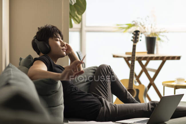 Молодой китаец сидит на диване с ноутбуком, слушает музыку в наушниках с закрытыми глазами и держит карандаш — стоковое фото