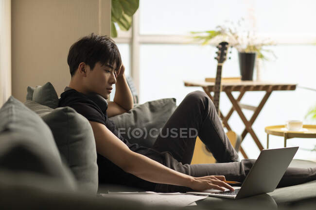 Jovem chinês homem usando laptop sentado no sofá — Fotografia de Stock