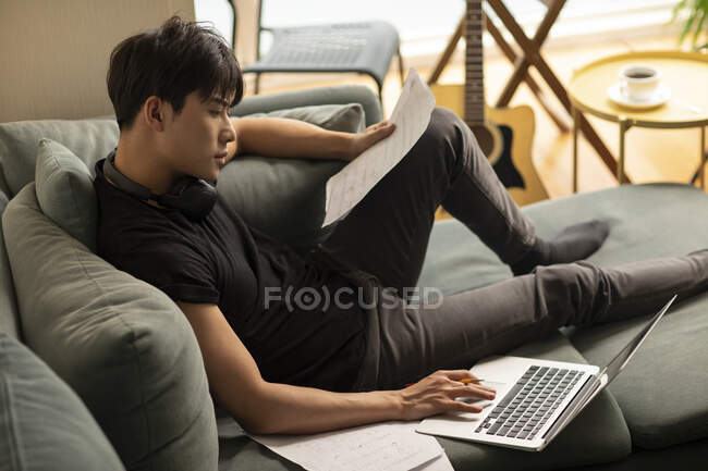 Hombre chino joven usando el ordenador portátil con papeles en el sofá - foto de stock