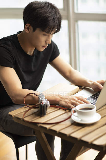 Hombre chino joven usando el ordenador portátil en la mesa con taza de café y cámara vintage - foto de stock