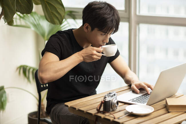 Jovem chinês homem beber café da xícara e usando laptop — Fotografia de Stock