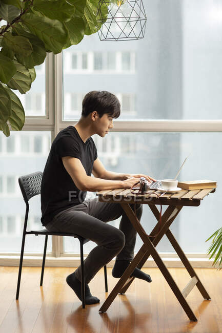 Jeune homme chinois utilisant un ordinateur portable à la table avec livre, tasse à café et appareil photo vintage — Photo de stock