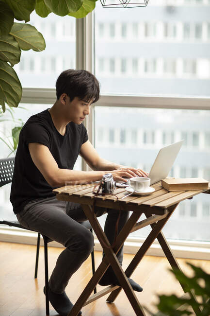 Hombre chino joven usando el ordenador portátil en la mesa con libro, taza de café y cámara vintage - foto de stock