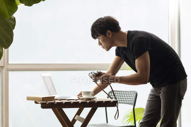 Молодой китаец, стоящий и держащий винтажную камеру во время использования ноутбука — стоковое фото