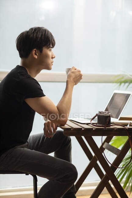 Jovem chinês homem sentado com xícara de café na mesa com laptop — Fotografia de Stock