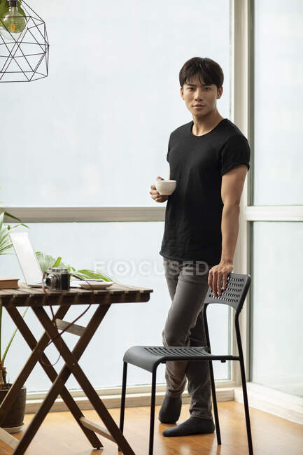 Young chinese man standing with coffee cup by table and looking at camera — Stock Photo