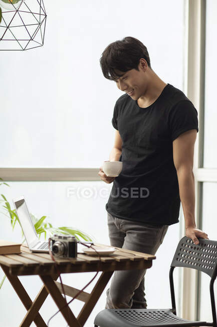 Giovane cinese con tazza di caffè guardando il computer portatile sul tavolo e sorridendo — Foto stock