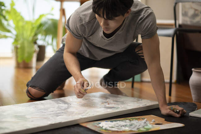 Young chinese man holding brush with canvas, paints on floor — Stock Photo