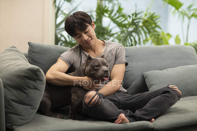 Молодий китайський чоловік обіймає собаку, сидячи на дивані — стокове фото