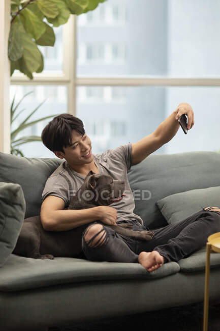 Young chinese man taking selfie with dog on couch and smiling — Stock Photo