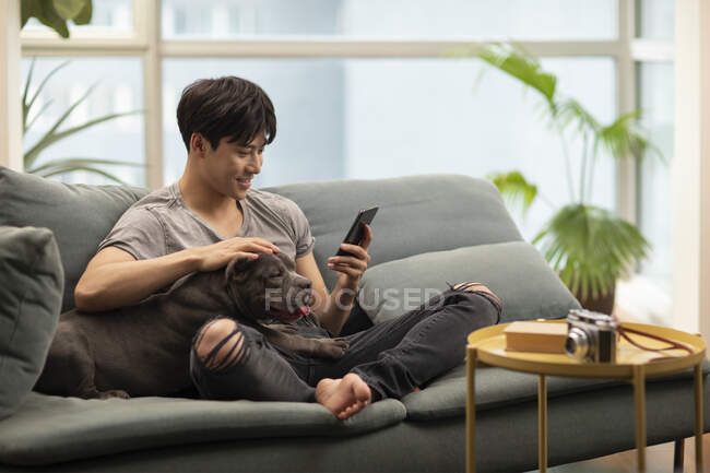 Hombre joven chino acariciando perro y sonriendo mientras mira el teléfono inteligente - foto de stock