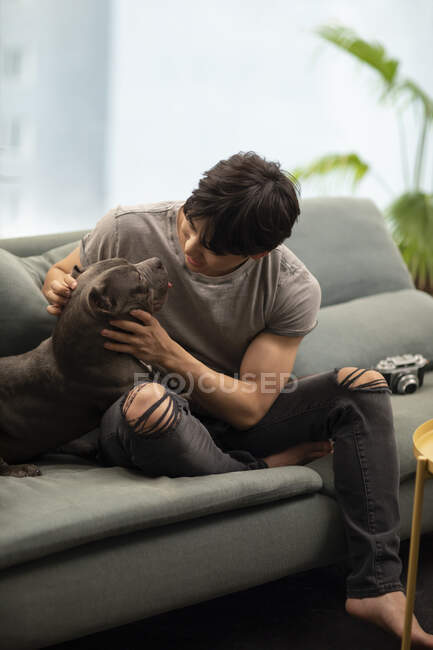 Молодой китаец ласкает собаку, сидя на диване — стоковое фото
