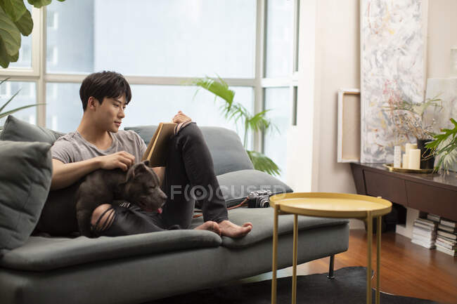 Junger Chinese mit Hund sitzt auf Couch und liest Buch — Stockfoto