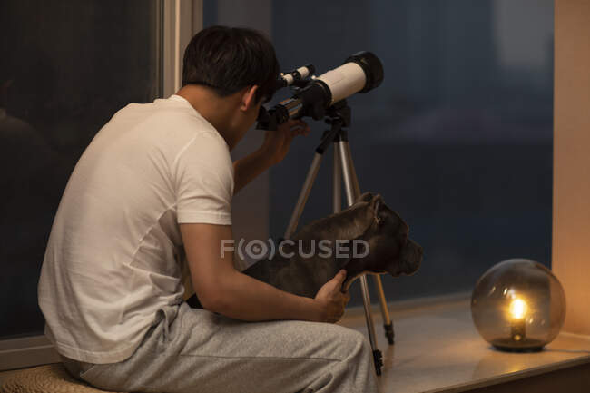 Jeune homme chinois assis avec chien et regardant à travers le télescope — Photo de stock