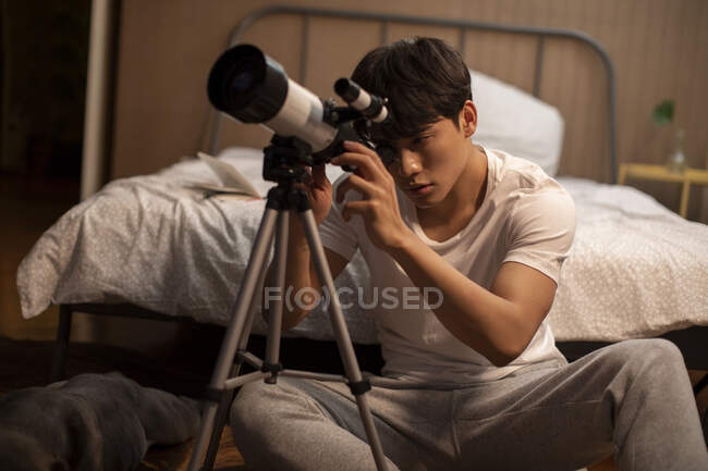 Young chinese man looking through telescope sitting beside bed — Stock Photo