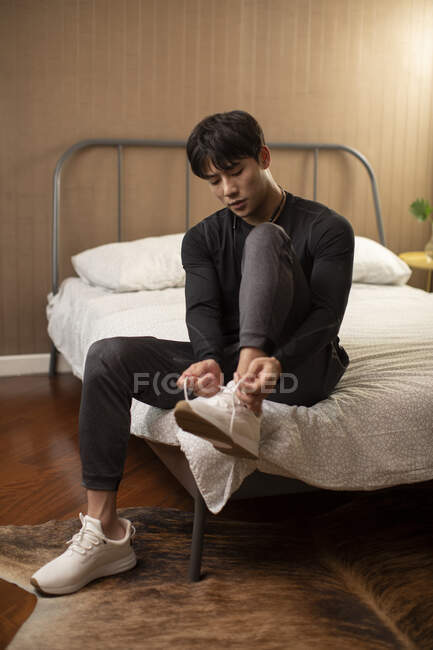 Jovem chinês homem amarrando atacadores em tênis enquanto sentado na cama — Fotografia de Stock