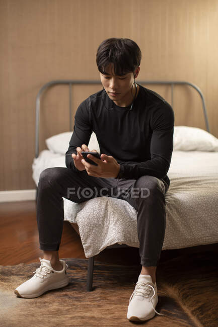 Giovane cinese con auricolari che guarda lo schermo dello smartphone mentre è seduto sul letto — Foto stock