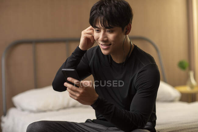 Улыбающийся китаец смотрит на экран смартфона и пользуется наушниками — стоковое фото