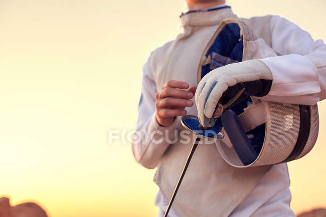 Esgrimista vestindo traje de esgrima branco e segurando sua máscara de esgrima e uma espada no fundo ensolarado — Fotografia de Stock