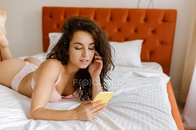 Primer plano de la joven caucásica bonita mujer en ropa interior descansando en casa en el dormitorio utilizando el teléfono móvil. Mujer atractiva en lencería acostada en la cama escribiendo en el teléfono inteligente mirando la pantalla del dispositivo - foto de stock