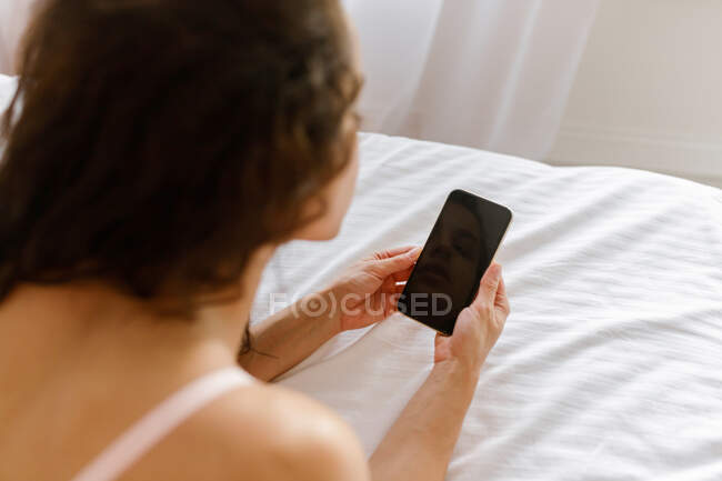Over shoulder view of young brunette woman in bra resting on bed indoors in the morning holding in hands smartphone looking at device screen. Close up of mobile phone with black display in female hand — Stock Photo