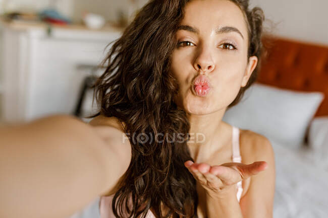 Close up. POV of Caucasian pretty young brunette woman indoor posing to smartphone camera taking selfie photos. Happy beautiful female in bedroom takes picture of herself giving air kiss — Stock Photo
